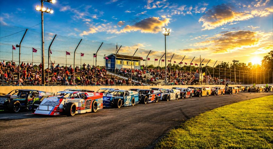 Cars lined up on the front stretch at sunset in front of the grandstands